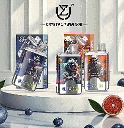 Assorted Crystal Twin 50K vape devices displayed on a white round platform, surrounded by blueberries and half a grapefruit, with branded packaging in the background.