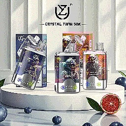 Assorted Crystal Twin 50K vape devices displayed on a white round platform, surrounded by blueberries and half a grapefruit, with branded packaging in the background.