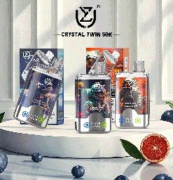 Assorted Crystal Twin 50K vape devices displayed on a white round platform, surrounded by blueberries and half a grapefruit, with branded packaging in the background.